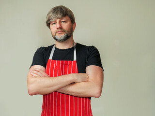 Portrait of professional butcher or fishmonger in black shirt and red and white classic traditional apron, light color background. Male model in his 40s with grey beard and hair. Hands crossed