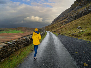 Teenager girl tourist in blue jeans and yellow hat and jacket walking on wet narrow road in a mountain. Travel and tourism. Sightseeing in Connemara, Ireland.