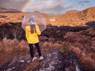 Teenager girl in yellow jacket and umbrella and enjoys stunning mountain view in Killarney area of Ireland. Travel and tourism. Outdoor activity. Model with long hair and slim body type.