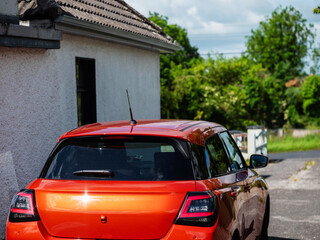 Small sport red car parked by a house with white walls, rural area in the background. Warm sunny day. Living in a country side concept. Commute and transportation theme.