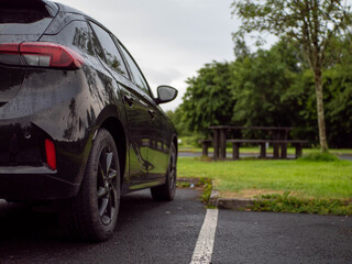 Black family car parked in a parking lot of a park with barbecue or dining area, selective focus. Nobody. Car surface is with rain drops. Outdoor activity. Travel and tourism.