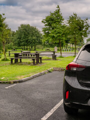 Black family car parked in a parking lot of a park with barbecue or dining area, selective focus. Nobody. Car surface is with rain drops. Outdoor activity. Travel and tourism.
