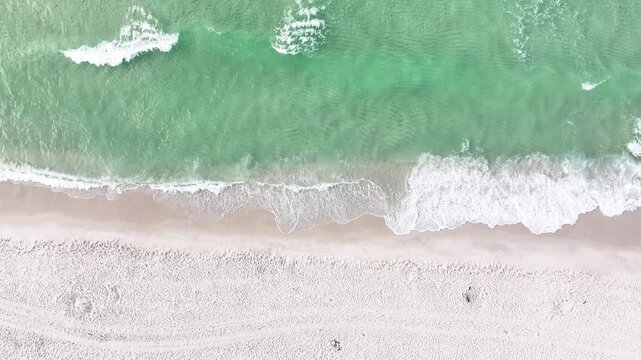Slow-motion video of a birds-eye-view of ocean waves crashing onto a sandy beach in Pensacola, Florida, on a beautiful sunny summer day
