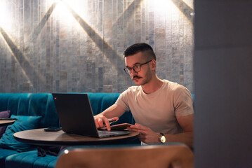 Focused young man with glasses writing in a notebook while working on a laptop in a modern cafe....