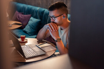 Focused young man wearing glasses and wireless earbuds, watching a webinar, having an online course on a laptop in a modern interior. 