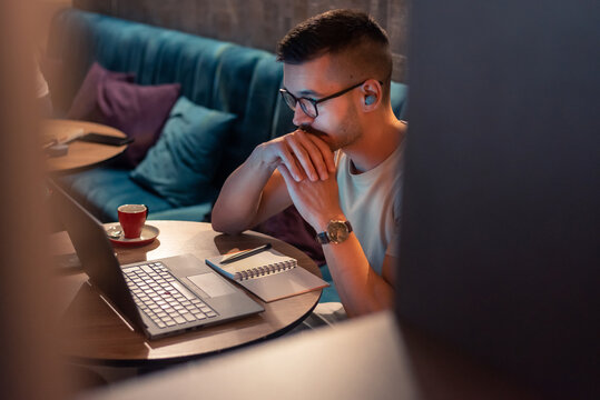 Focused young man wearing glasses and wireless earbuds, watching a webinar, having an online course on a laptop in a modern interior. 