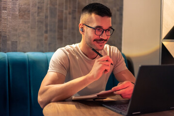 A young happy man wearing glasses and wireless earbuds, watching a webinar, having a video call...