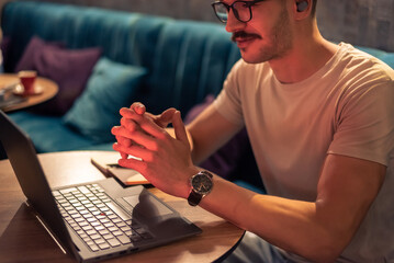 A happy young man working in a modern cafe, wearing glasses and wireless earbuds, having a video call with clients on a laptop, concept of remote work, freelancer lifestyle or online learning.