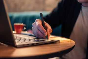 Close cropped image of a business man writing in a sticky note while working on a laptop or a male...
