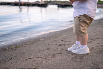 Toddler in white sneakers explores the beach. Boy in beige shorts runs barefoot in the sand.