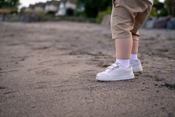 Toddler in white sneakers explores the beach. Boy in beige shorts runs barefoot in the sand.