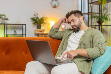 Overtired Caucasian young man with brown hair bored using laptop while sitting on sofa at home....