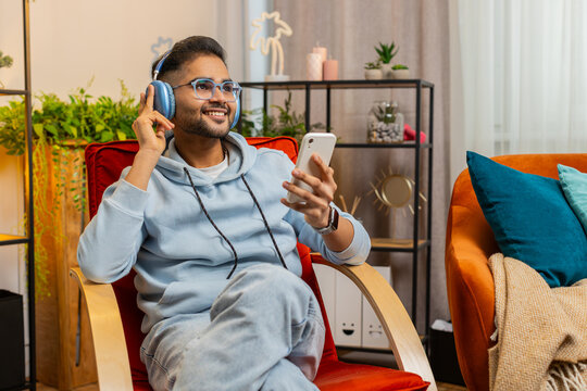 Happy relaxed overjoyed Indian Arabian man in wireless headphones dancing on chair at home choosing listening favorite energetic disco pop music in smartphone entertaining. People weekend activities