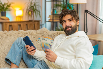 Planning budget. Rich happy Indian man using smartphone calculator app count euro cash, calculate...