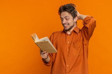 Happy relaxed smiling Caucasian man reading funny interesting fairytale story book, leisure hobby, knowledge wisdom, education, learning, study, wow. Young guy isolated on orange background indoors