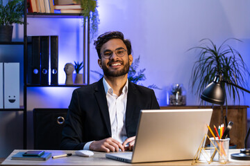 Portrait of happy smiling bearded Indian businessman at modern home office workplace desk looking at camera. Male Hindu Arabian in formal suit. Handsome man remote distant online working on laptop
