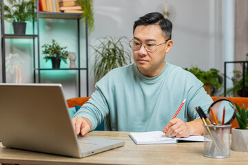 Asian man freelancer making notes on notebook at home, use laptop, reading information prepare...