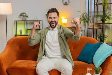 Happy Caucasian young man watching TV shouting, celebrating success, winning, goal achievement good...