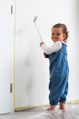 Cute little curly haired girl painting a white wall with a roller, wearing denim overalls and a...