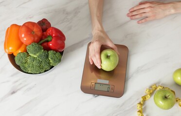 Woman weighting apple on kitchen scale at white marble table, top view