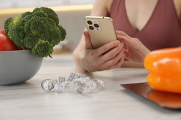 Woman using smartphone at white marble table with vegetables, kitchen scale and measuring tape...