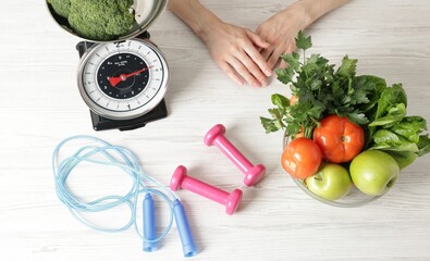 Woman at light wooden table with healthy food, kitchen scale, skipping rope and dumbbells, top view