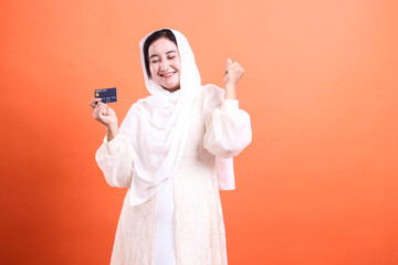 Excited Muslim Woman Celebrating with Credit Card Success, showcasing financial empowerment and joy isolated on a orange background.