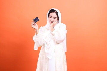 Worried Muslim Woman with Credit Card: Financial Concerns, isolated on a orange background,...