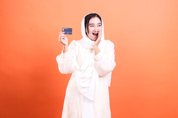Excited Muslim Woman with Credit Card Celebrating Financial Win, isolated on an orange background, full of joy and modern financial opportunities.