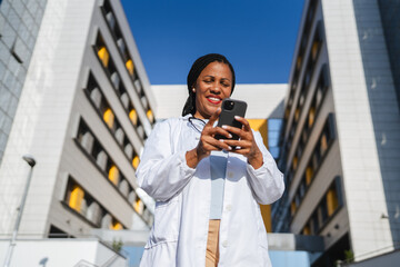 african american woman doctor on break use cellphone in front hospital