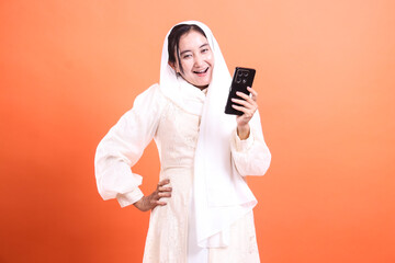 Joyful Muslim Woman with Smartphone: Smiling and holding a mobile phone while looking at the camera happily isolated on a orange background.
