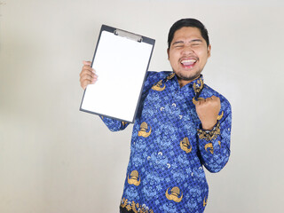 Indonesian civil servant man shows white paper attached to clipboard with excited expression....