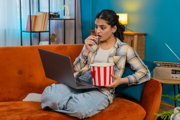 Indian young woman at home sitting on sofa, watching movie on laptop, laughing loudly with lively...