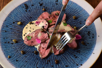 Closeup person's hands using fork and knife to cut meat of Vitello Tonnato dish on blue plate