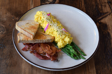 Breakfast plate with scrambled eggs, bacon, steamed asparagus and toasted bread on wooden table