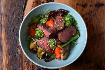 Bowl with warm sliced roast beef and grilled vegetables salad on wooden table