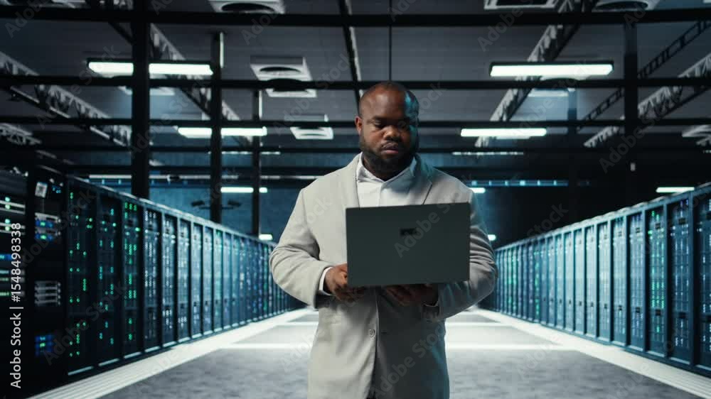 Poster engineer walking in server room backing up storage arrays using software on laptop. african american