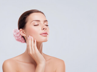 Woman with flawless skin gently touching her face while wearing a flower in her hair against a neutral background