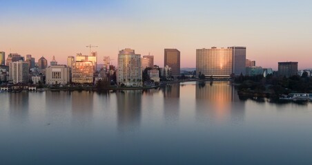 Fototapeta premium Lake Merritt reflects the Oakland, California, USA skyline at sunset. The buildings mirror in the water, creating a serene urban landscape.
