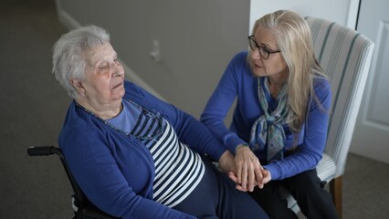 An elderly woman in a wheelchair receives loving support from her daughter during a visit. They...