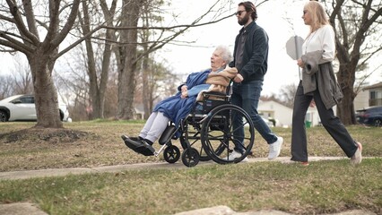 Obraz premium Elderly mother in a wheelchair is taken for a walk by her daughter and grandson on a sunny spring day, sharing smiles and happiness outdoors together.