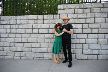 woman in green dress in hat and man in hat are near brick wall,
