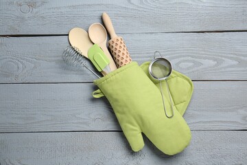 Different kitchen utensils in oven mitten on light grey wooden table, top view