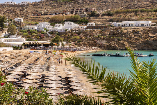 Wide angle view of beach club on Greek islands with lots of umbrellas and seating against a rocky landscape and blue sea.