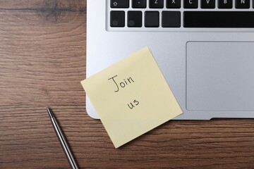 Paper note with words Join Us, laptop and pen on wooden table, flat lay