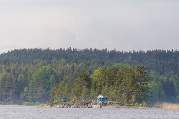 A small blue tent stands on a rocky forested island surrounded by calm lake waters and dense pine...