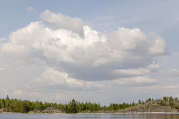 Dramatic cumulus clouds hover above a serene forested shoreline with rocky terrain and calm...