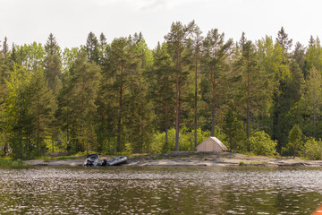 A beige canvas tent and two inflatable boats rest on a rocky shoreline in a forest, reflecting...