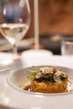Fine dining entree of tartare garnished with greens and shaved truffles with a glass of wine in the background.  