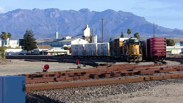 A locomotive and freight cars park in a rail yard in an industrial zone of Oxnard as cars whiz by against a background of the Santa Monica Mountains.
